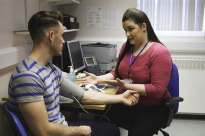 A horizontal image of a young man sitting getting his blood pressure taking by a female doctor. They are in a typically British doctor's practice chatting together, while she performs the medical examination.