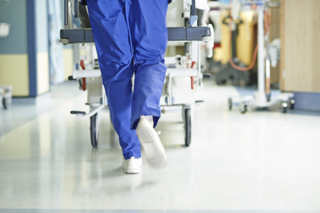 legs of a healthcare worker in a blue uniform, running down a hospital corridor and pushing a trolley