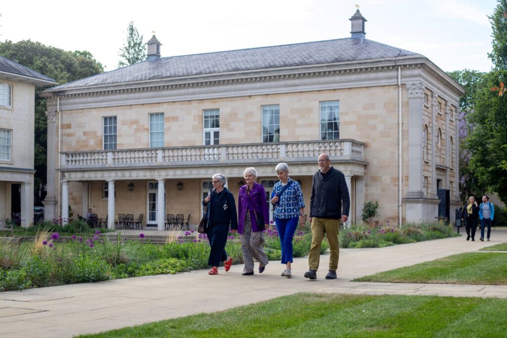 Four adults walking along a path in a landscaped garden beside a large classical stone building with columns and a balcony.