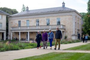 Four adults walking along a path in a landscaped garden beside a large classical stone building with columns and a balcony.