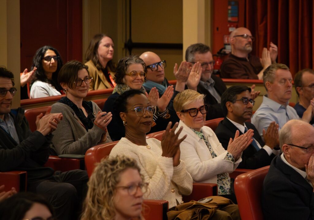 Audience seated in a lecture hall applauding, with rows of people facing forward.