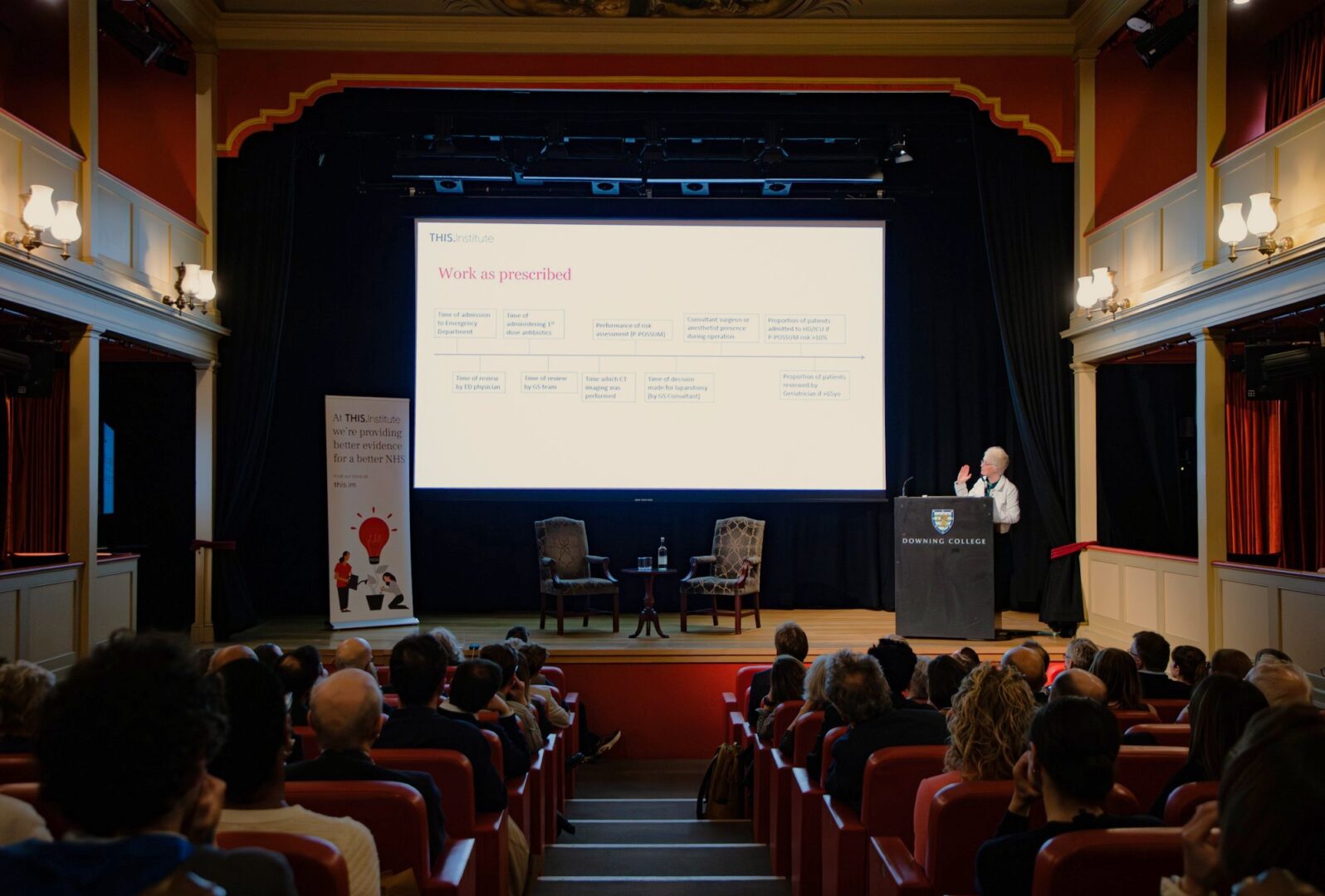 Mary Dixon-Woods stands at a lectern on a stage in a theatre-style auditorium, presenting to a seated audience. Behind the speaker, a large projected slide shows a flow diagram. Two empty chairs and a small table with water are arranged on stage. A banner to the left reads “At THIS Institute we’re providing better evidence for a better NHS.” The room features red seating, warm lighting, and decorative wall lamps.