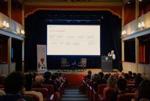 Mary Dixon-Woods stands at a lectern on a stage in a theatre-style auditorium, presenting to a seated audience. Behind the speaker, a large projected slide shows a flow diagram. Two empty chairs and a small table with water are arranged on stage. A banner to the left reads “At THIS Institute we’re providing better evidence for a better NHS.” The room features red seating, warm lighting, and decorative wall lamps.