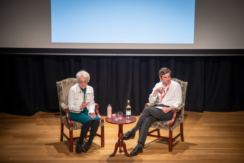 Two speakers seated on stage in armchairs during a discussion, with a small table between them holding water bottles and glasses beneath a large projection screen.