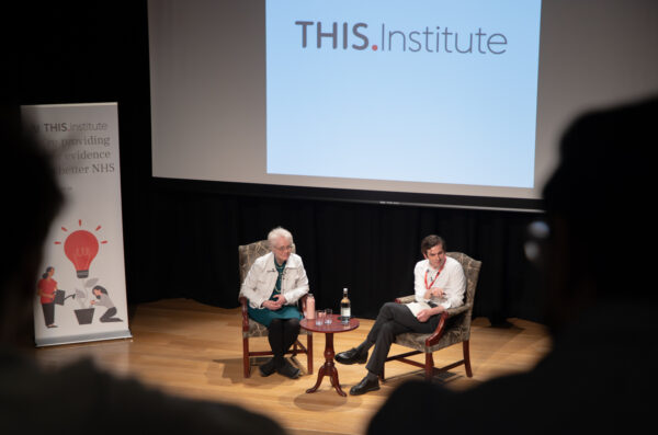 Two speakers sit on armchairs on a stage during a Q&A session at 2026 THIS Institute Annual Lecture, seen from the audience. A large screen behind them displays the text “THIS Institute.” A small table between the speakers holds a bottle of water and two glasses. A standing banner to the left reads, “At THIS Institute we’re providing better evidence for a better NHS,” alongside an illustration of a lightbulb and figures. The audience is visible in the foreground in soft focus.