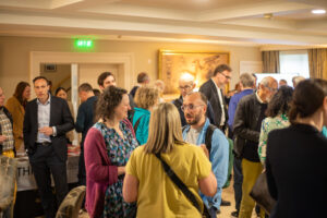 Crowd of people in a bright reception room networking and talking in small groups, with a display table and wall art in the background.