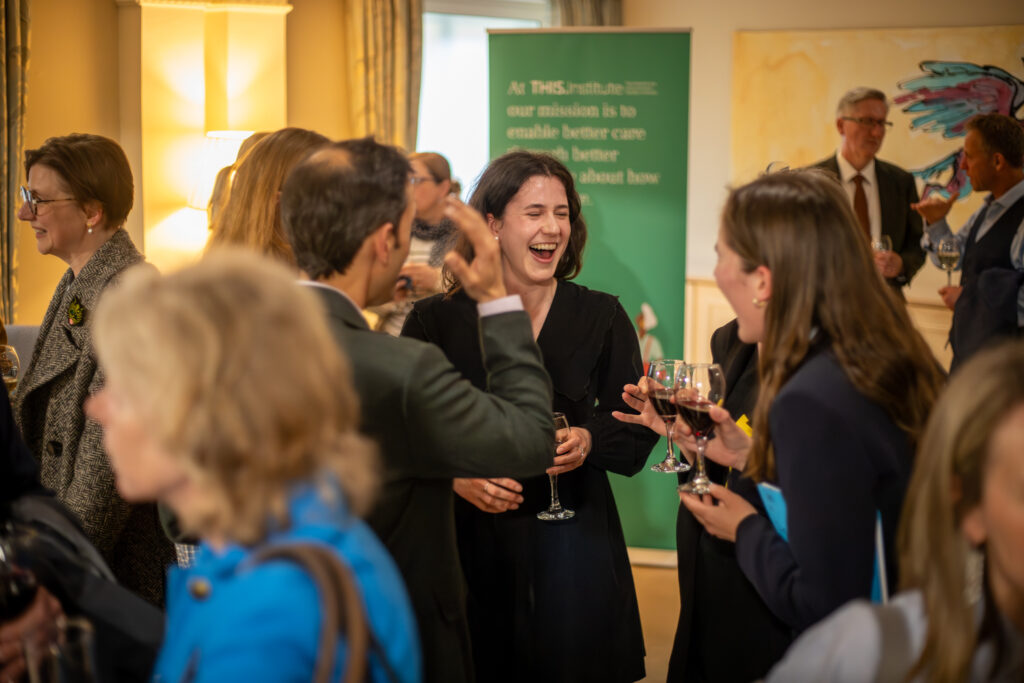 THIS Institute's annual lecture attendees chatting and holding drinks, gathered near a green banner that reads, “At THIS Institute our mission is to enable better care.”