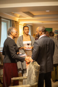 Two people in conversation at an indoor networking event, standing close together while others talk around them; a branded THIS Institute display and table are visible in the background.