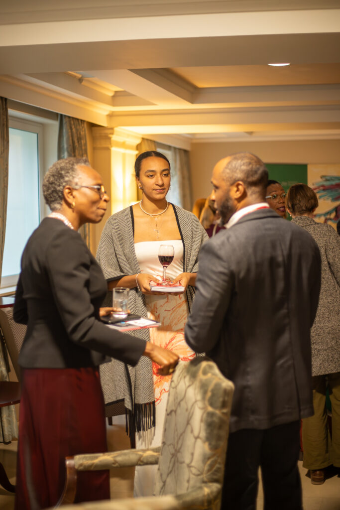 Two people in conversation at an indoor networking event, standing close together while others talk around them; a branded THIS Institute display and table are visible in the background.