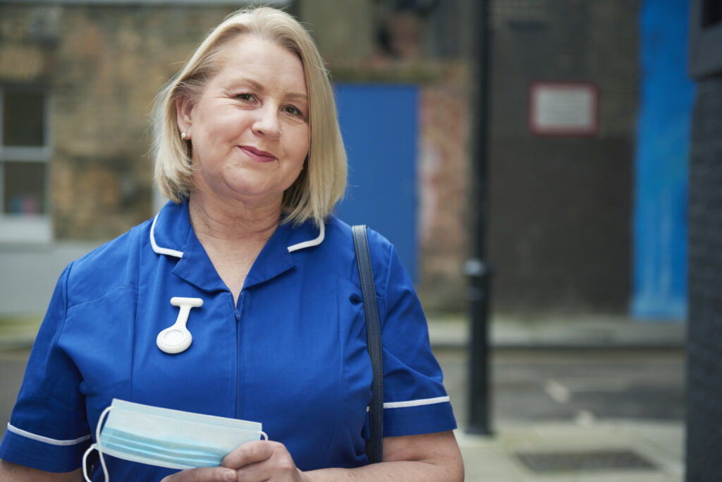 Female nurse in blue uniform standing in front of a hospital building holding clipboard